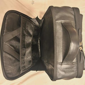 May include: Black leather backpack with multiple interior compartments and a handle. The backpack is open, revealing several pockets and elastic straps. The exterior features a zipper closure and a textured leather finish.