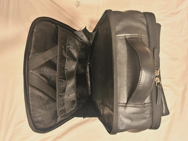 May include: Black leather backpack with multiple interior compartments and a handle. The backpack is open, revealing several pockets and elastic straps. The exterior features a zipper closure and a textured leather finish.