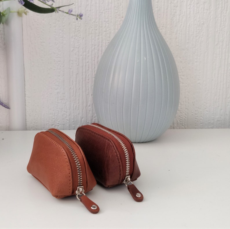 May include: Two small, brown leather coin purses with zipper closures. The purses are lying on a white surface.