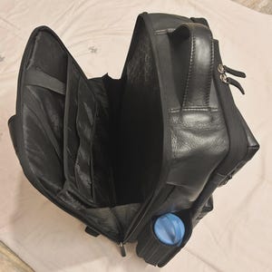 May include: Black leather backpack with multiple compartments, including a side pocket holding a blue water bottle. The bag has a top handle and a zippered closure. The interior features several organizational pockets.