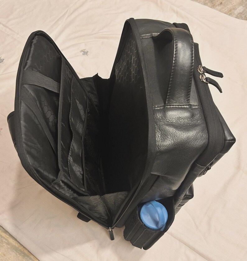 May include: Black leather backpack with multiple compartments, including a side pocket holding a blue water bottle. The bag has a top handle and a zippered closure. The interior features several organizational pockets.