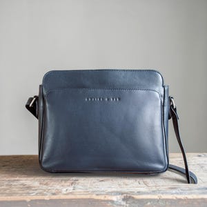 May include: A dark navy blue leather crossbody bag with a rectangular shape, featuring a smooth front panel, a top zipper closure, and silver-toned metal hardware. The brand name "LEATHER & TAN" is embossed in small capital letters on the front.