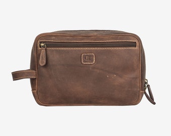 May include: A brown leather toiletry bag with a rectangular shape. The bag features a top zipper, a front zipper pocket, and a small handle on the side. The stitching is a light tan color, and the bag has a distressed look.