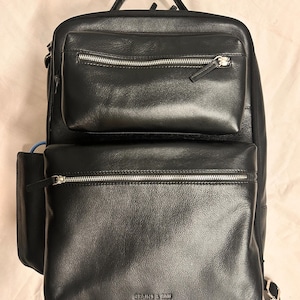 May include: Black leather backpack with multiple zippered compartments and a top handle. The backpack features silver zippers and the brand name "GRAINS & TAN" is visible on the front. The bag has a structured design.