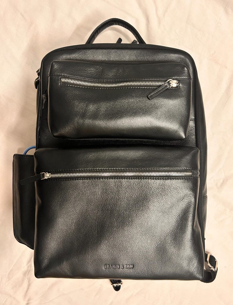 May include: Black leather backpack with multiple zippered compartments and a top handle. The backpack features silver zippers and the brand name "GRAINS & TAN" is visible on the front. The bag has a structured design.