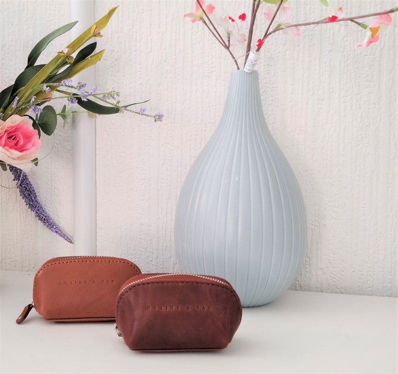 May include: Two small brown leather pouches with zipper closures. The pouches have the text "Leather & Co" embossed on them. The pouches are sitting on a white surface next to a light blue vase with pink flowers.