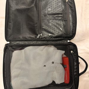 May include: Open black carry-on suitcase with a mesh compartment containing a gray garment and a red cylindrical object. The top section has multiple zippered compartments and a honeycomb pattern lining.