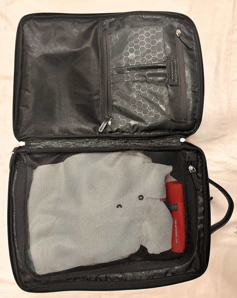 May include: Open black carry-on suitcase with a mesh compartment containing a gray garment and a red cylindrical object. The top section has multiple zippered compartments and a honeycomb pattern lining.