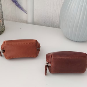 May include: Two small brown leather zippered pouches. The pouch on the left is a lighter brown and the pouch on the right is a darker brown.