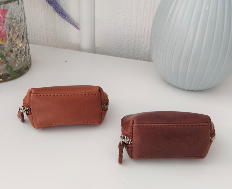 May include: Two small brown leather zippered pouches. The pouch on the left is a lighter brown and the pouch on the right is a darker brown.
