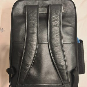 May include: Black leather backpack with padded shoulder straps and a top handle. The backpack has a rectangular shape with a zippered main compartment and a side pocket. The leather has a textured appearance.