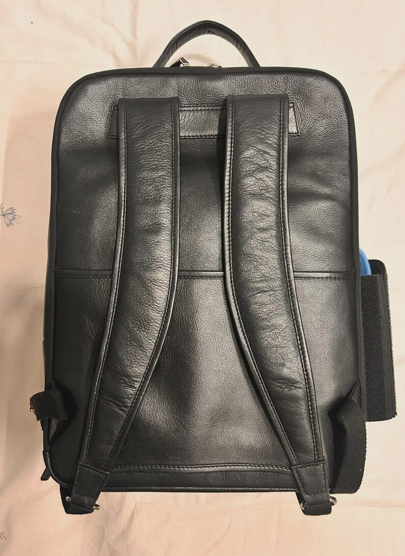 May include: Black leather backpack with padded shoulder straps and a top handle. The backpack has a rectangular shape with a zippered main compartment and a side pocket. The leather has a textured appearance.