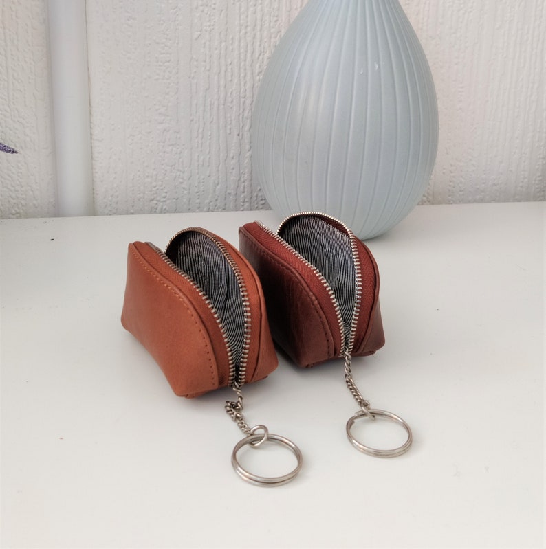 May include: Two brown leather keychains with a zipper closure and a silver key ring. The keychains are open and the inside lining is black and white striped.
