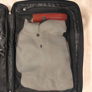 May include: Open black travel suitcase with a handle, zipper, and mesh compartments. Inside, a gray button-down shirt is visible, along with a red cylindrical object. The suitcase is partially open, revealing its interior organization.