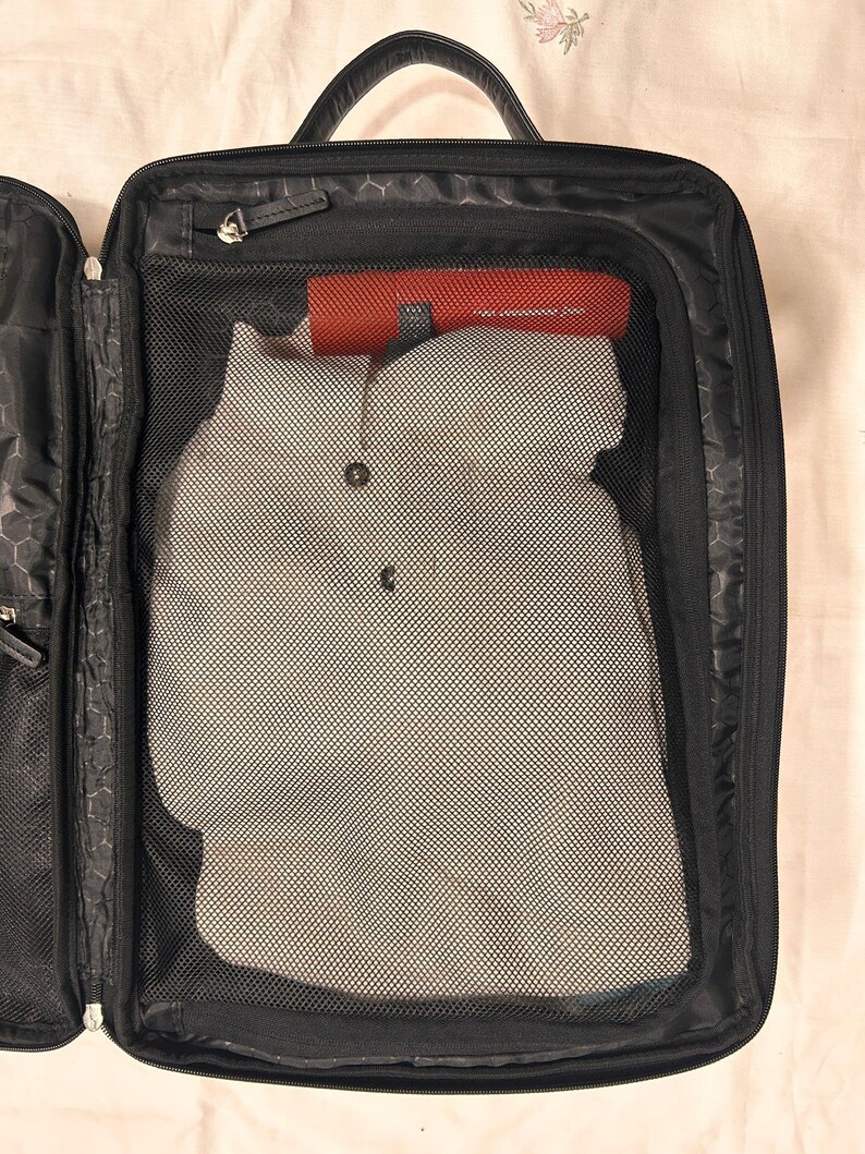 May include: Open black travel suitcase with a handle, zipper, and mesh compartments. Inside, a gray button-down shirt is visible, along with a red cylindrical object. The suitcase is partially open, revealing its interior organization.