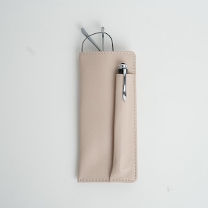 Könnte beinhalten: Ein hellbeiger Leder-Brillenetui mit Stifthalter. Das Etui hat einen genähten Rand und eine Klappe, die mit einem Druckknopf geschlossen wird.