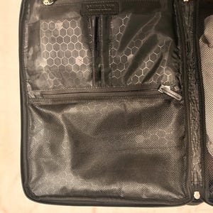 May include: Black travel organizer with multiple compartments and zippers. The interior features a honeycomb pattern and a label that reads "GRAIN & TAN GENUINE LEATHER". The organizer is open, displaying its interior pockets and mesh sections.