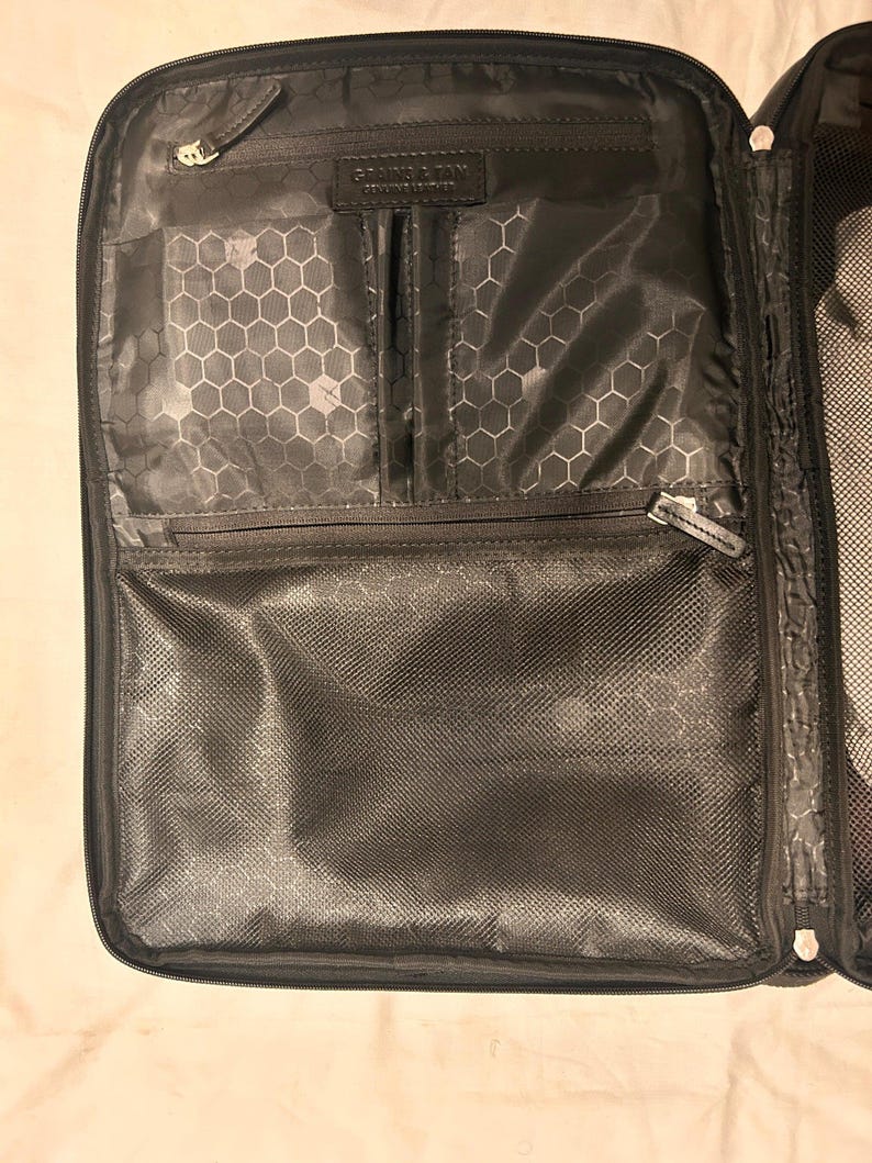 May include: Black travel organizer with multiple compartments and zippers. The interior features a honeycomb pattern and a label that reads "GRAIN & TAN GENUINE LEATHER". The organizer is open, displaying its interior pockets and mesh sections.