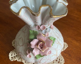 May include: A white porcelain vase with a ruffled top and gold trim. The vase is decorated with pink and white flowers and a pink rose with gold accents. The vase is sitting on a white crocheted doily.