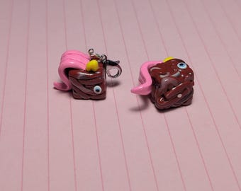 May include: Two square-shaped brown charms with a glossy finish, each featuring a single blue eye, a small yellow horn, and a flowing pink hair-like attachment. One charm includes a silver-toned metal lobster clasp attached to the top corner.