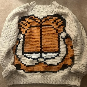 May include: A beige crochet sweater featuring a large orange and black Garfield face on the front. The sweater has long sleeves and a crew neck. The design is a pixelated style, giving it a retro look.