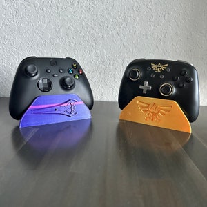 May include: Two black video game controllers with different colored stands. The controller on the left has a purple stand with a stylized design. The controller on the right has a gold stand with a stylized design.
