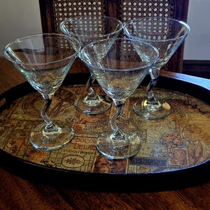 May include: Four clear glass martini glasses with a unique, twisted stem design. The glasses sit on a decorative, oval tray with a vintage map design. The tray has a black rim and is placed on a wooden surface.