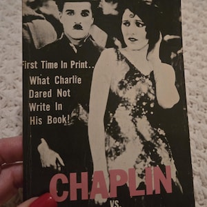 May include: Vintage paperback book cover featuring Charlie Chaplin and a woman. The cover is black and white with the title "CHAPLIN vs. CHAPLIN" in pink. Text includes "First Time In Print... What Charlie Dared Not Write In His Book!"
