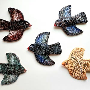 May include: Five ceramic bird figurines in flight, each with a unique color scheme. The birds feature textured surfaces and intricate circular patterns. Colors include brown, blue, green, and tan, with red beaks. The birds are arranged on a white background.