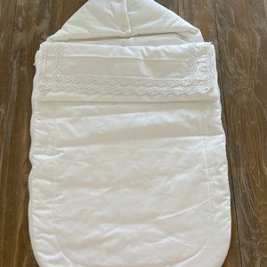 May include: White baby sleeping bag with a lace trim and a zipper closure.