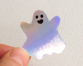 May include: A holographic ghost sticker with a smiling face. The sticker has a pearlescent sheen, reflecting various colors. The ghost has two small black eyes and a black open mouth. The sticker is held by a person's fingers.