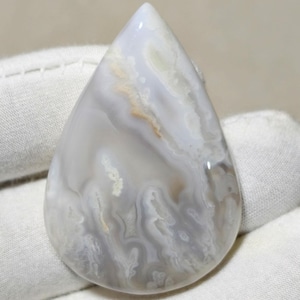 Può includere: Una gemma a forma di goccia lucidata. La pietra presenta un motivo a vortice di grigio, bianco e sfumature di beige. Il design evoca l'acqua che scorre o le nuvole. La gemma è tenuta in un guanto bianco.