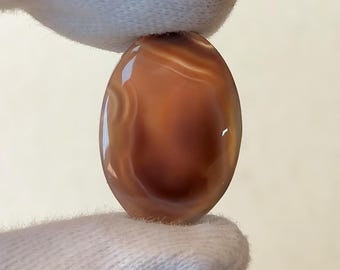 Pode incluir: Um cabochão de ágata liso de forma oval com bandas marrons e brancas. A pedra tem um acabamento polido e é translúcida.