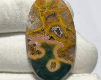 May include: Oval-shaped gemstone with a unique pattern. The stone features a blend of colors, including yellow, green, pink, and white. The yellow and green hues dominate the top and bottom, while pink and white accents add contrast. The stone's surface has a polished appearance.