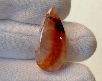 May include: A teardrop-shaped gemstone with a polished surface. The stone features a blend of colors, including translucent white, orange, and dark brown. The gemstone is held by a gloved hand.