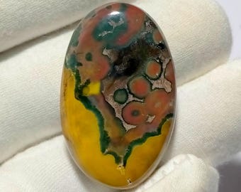 May include: An oval-shaped gemstone with a polished surface. The stone features a vibrant mix of colors, including yellow, green, red, and brown, creating a unique pattern. The gemstone is held in a white fabric.