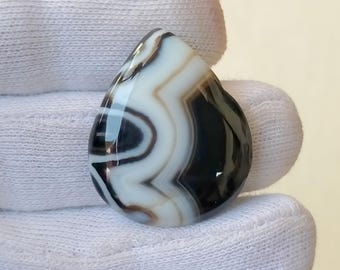 May include: A teardrop-shaped gemstone with a polished surface. The stone features a striking pattern of black, white, and brown bands, creating a unique, swirling design. The gemstone is held against a white background.