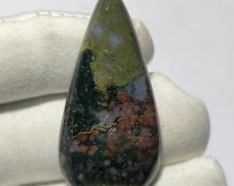 May include: A teardrop-shaped gemstone with a polished surface. The stone features a blend of colors, including green, red, black, and yellow, creating a unique pattern. The gemstone is held against a white background.