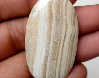 Puede incluir: Una piedra preciosa ovalada en cabujón blanca y marrón con un patrón de bandas. La piedra es lisa y pulida.