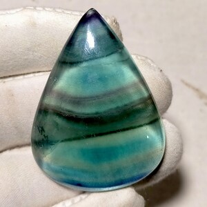 May include: A polished, teardrop-shaped gemstone. It displays bands of teal, blue, and purple. The translucent stone is held by a gloved hand, highlighting its natural patterns and colours. The gemstone is a beautiful example of natural artistry.