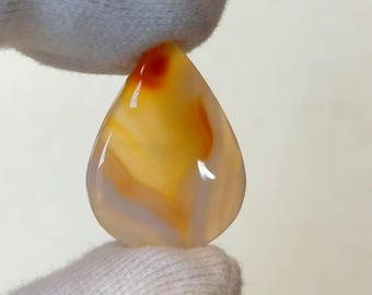 May include: A teardrop-shaped agate cabochon with white, orange, and red banding. The stone is translucent and has a smooth, polished surface.