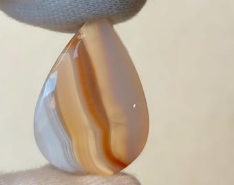 May include: A teardrop-shaped agate cabochon with white, orange, and brown banding.
