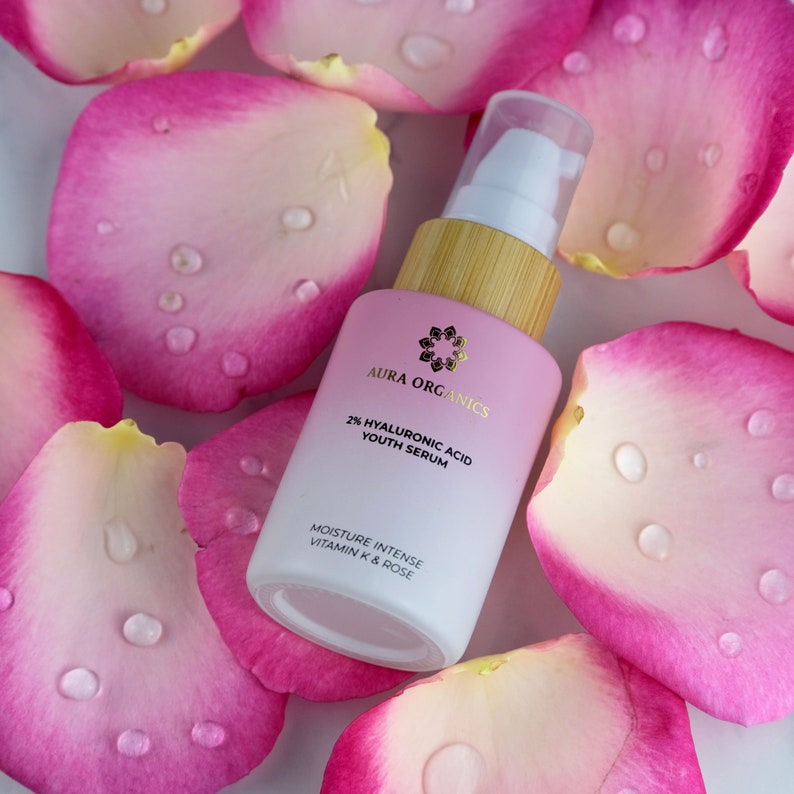 May include: A white bottle with a gold label and a bamboo lid. The label reads "Aura Organics 2% Hyaluronic Acid Youth Serum Moisture Intense Vitamin K & Rose". The bottle is surrounded by pink rose petals.