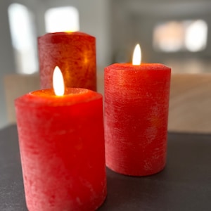 May include: Three red pillar candles with a textured surface are lit and burning. The candles are on a dark gray surface.