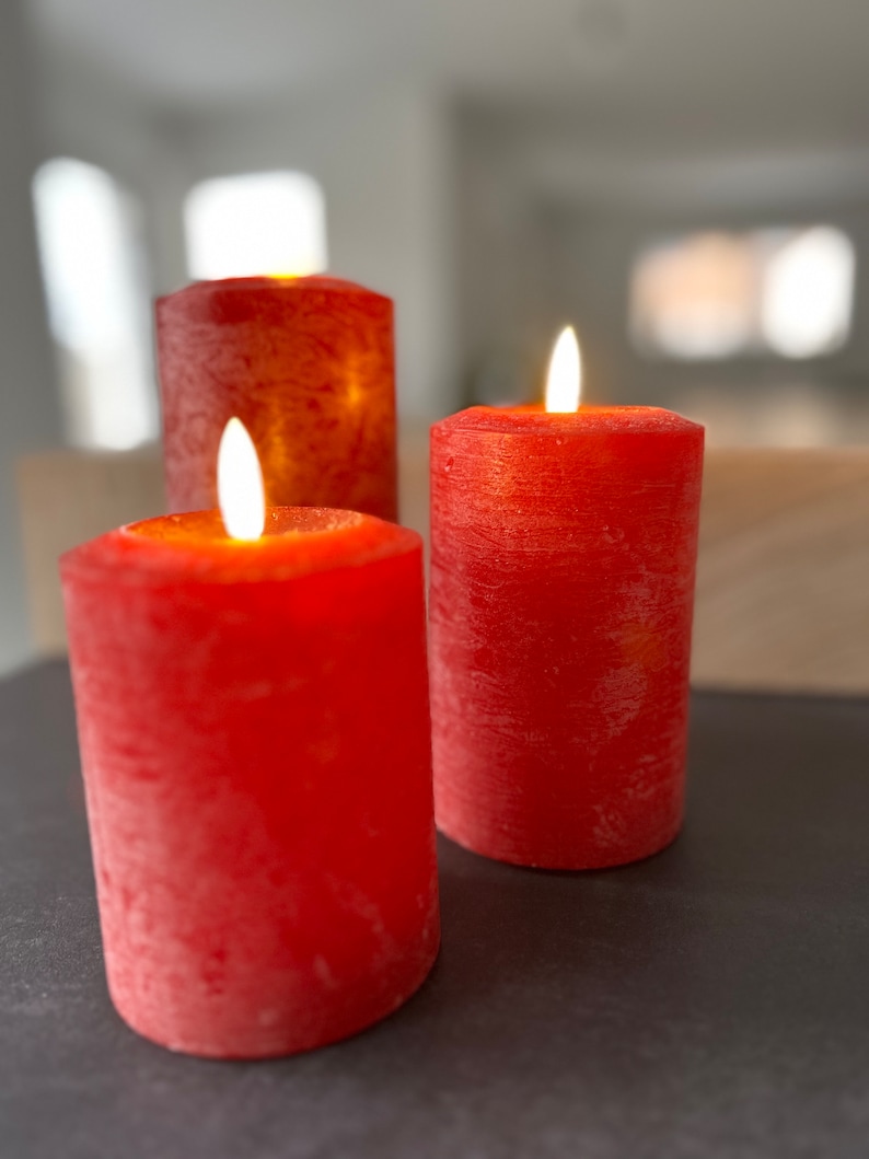 May include: Three red pillar candles with a textured surface are lit and burning. The candles are on a dark gray surface.