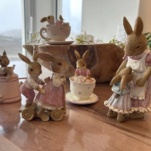 Könnte beinhalten: Eine Sammlung bezaubernder Hasenfiguren in verschiedenen Posen. Einige Hasen sitzen in Teetassen, andere schieben einen Wagen, und einer hält einen kleineren Hasen. Die Figuren sind in sanften Pastellfarben bemalt, was eine skurrile Szene schafft.