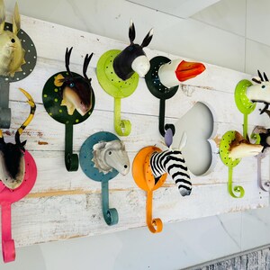Könnte beinhalten: Ein weißes Holzbrett mit zehn Wandhaken aus Metall, die jeweils mit einer bunten Tierkopf-Figur versehen sind. Die Tierköpfe umfassen einen Hasen, einen Hirsch, einen Esel, eine Gans, ein Schaf, ein Zebra, eine Ziege und weitere Arten.