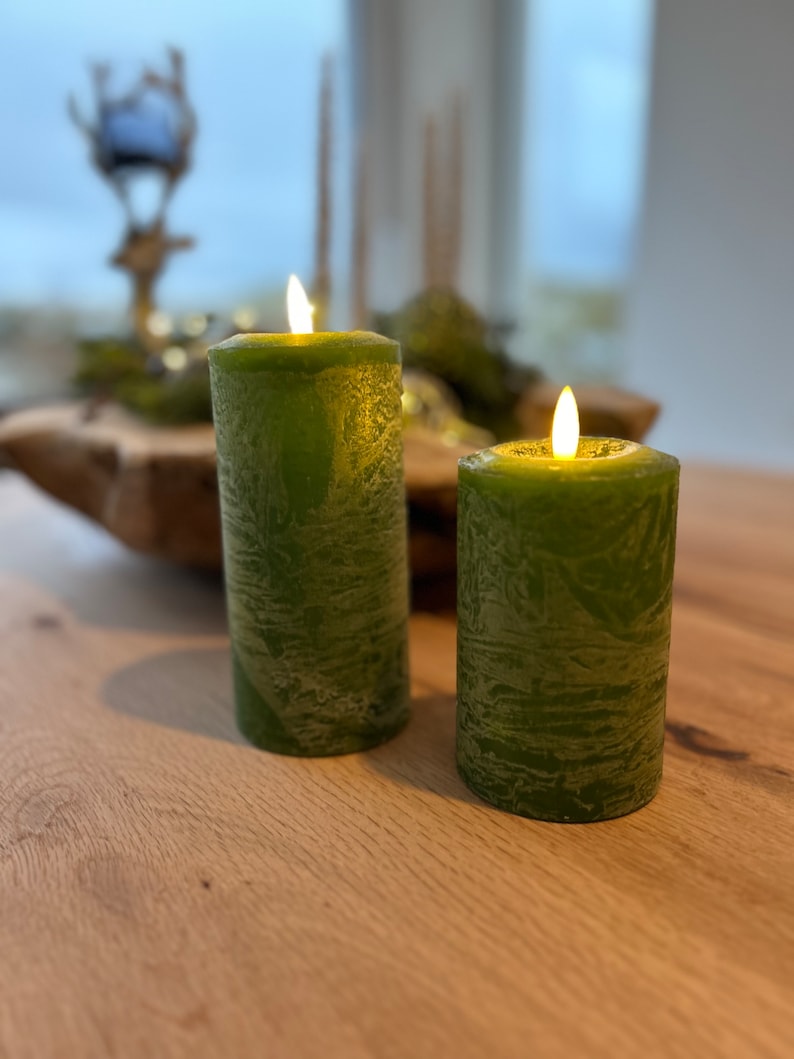 May include: Two green pillar candles with a textured surface are lit and sitting on a wooden table.