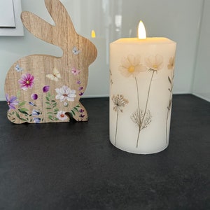 May include: A white pillar candle with dried flowers and a flickering flame. The candle is on a black surface next to a wooden rabbit cutout with floral designs.