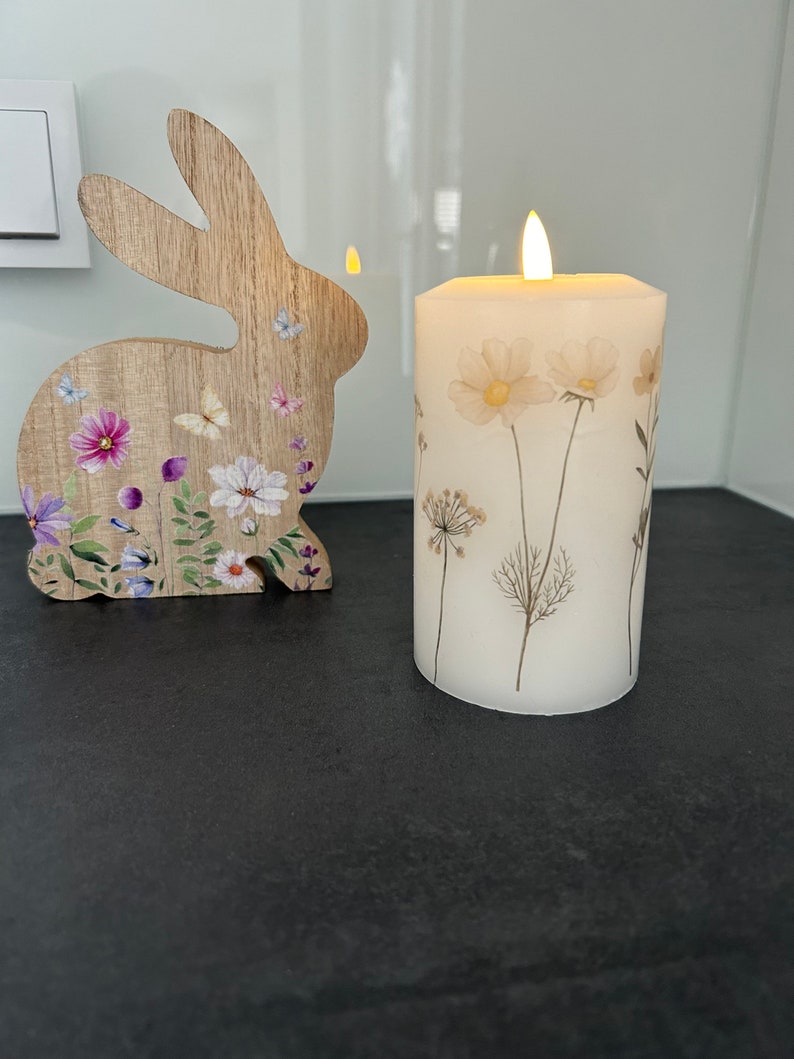 May include: A white pillar candle with dried flowers and a flickering flame. The candle is on a black surface next to a wooden rabbit cutout with floral designs.